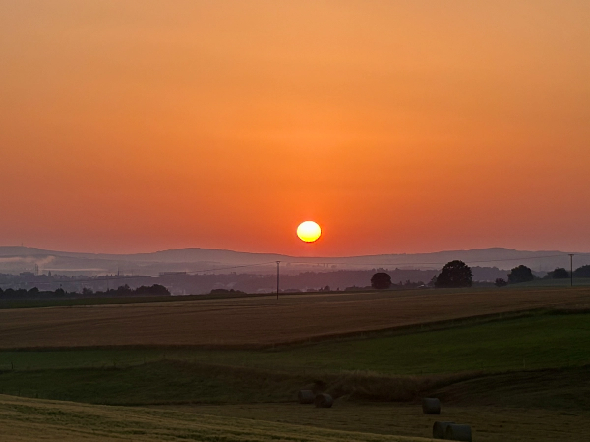 Sonnenuntergang über Feldern und sanften Hügeln, der Himmel leuchtet orange-rot.