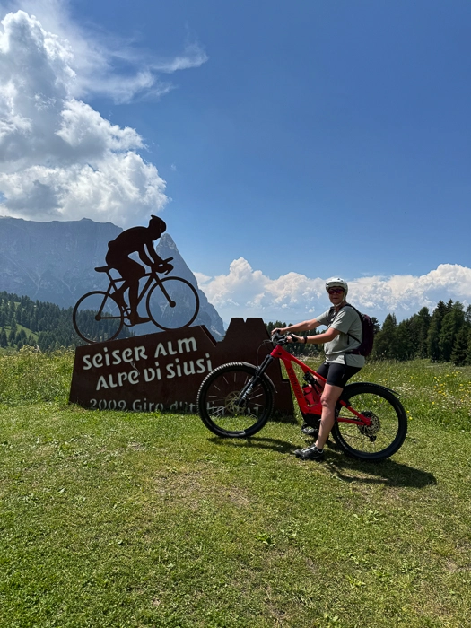 Eine Person steht mit einem roten Mountainbike vor dem Schild „Seiser Alm“ vor einer Bergkulisse unter blauem Himmel.