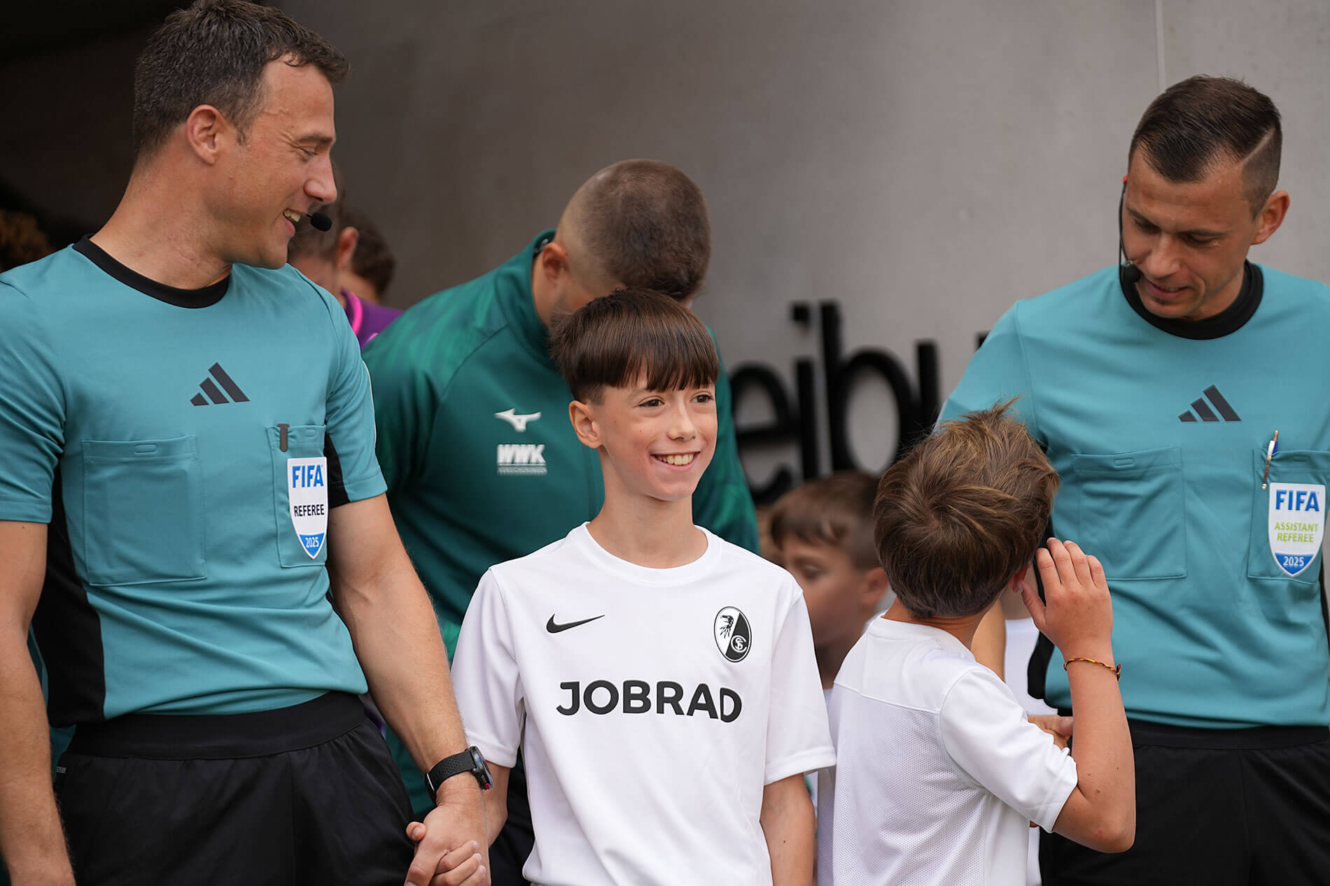 Schiri-Team mit Einlaufkindern. Ein Einlaufkind mit weißem Trikot mit JOBRAD-Aufdruck freut sich mächtig.